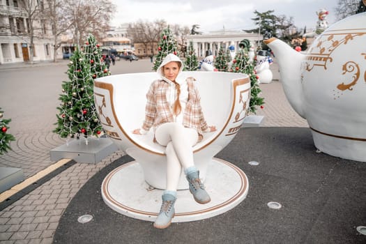 Woman Christmas Square. She sits in a large white cup, dressed in a light suit. With trees decorated with Christmas tinsel in the background.