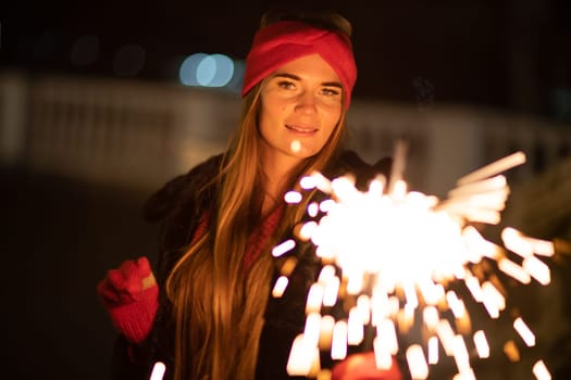 Woman holding sparkler night while celebrating Christmas outside. Dressed in a fur coat and a red headband. Blurred christmas decorations in the background. Selective focus.