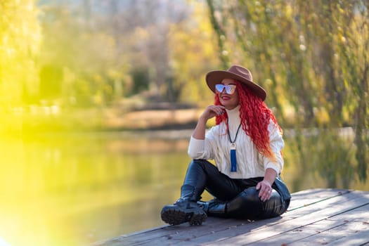 Autumn lake woman. In autumn, she sits by the pond on a wooden pier and admires nature with red hair and a hat. Tourism concept, weekend outside the city