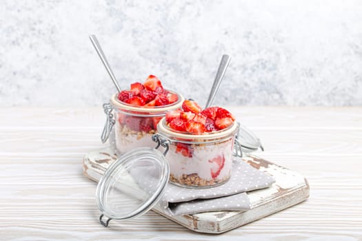 Parfait with Fresh Strawberries, Yoghurt and Crunchy Granola in Transparent Glass Mason Jars on White Rustic Wooden Background from Angle View, Healthy Breakfast or Light Summer Fruit Dessert