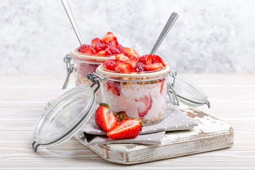 Parfait with Fresh Strawberries, Yoghurt and Crunchy Granola in Transparent Glass Mason Jars on White Rustic Wooden Background from Angle View, Healthy Breakfast or Light Summer Fruit Dessert