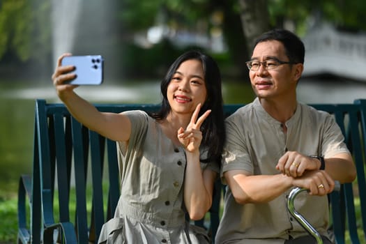 Happy smiling young woman with her father sitting on a wooden bench at public park making selfie on smartphone.