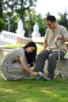 Caring young adult daughter helping her father tying shoelaces while relaxing in the public park.