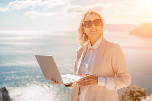 Freelance women sea. She is working on the computer. Good looking middle aged woman typing on a laptop keyboard outdoors with a beautiful sea view. The concept of remote work