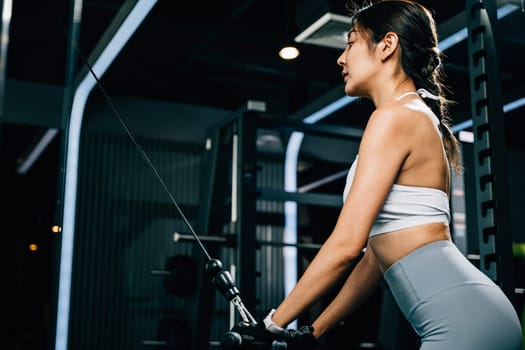 Fit Asian woman pulling down weights on a cable machine at the gym, training her biceps and building muscle strength. fitness center health workout concept