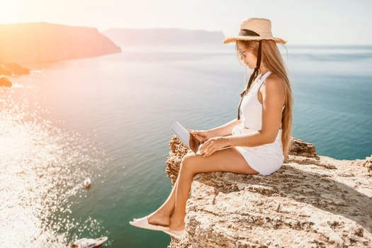 Successful business woman in yellow hat working on laptop by the sea. Pretty lady typing on computer at summer day outdoors. Freelance, travel and holidays concept.