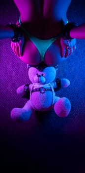 sexy buttocks of a girl in panties and hands behind her back in handcuffs , in an adult BDSM sex game and a teddy bear toy in the neon light