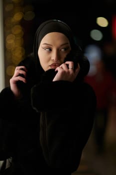 Young Muslim woman walking on urban city street on a cold winter night wearing hijab scarf veil a fashionable coat with bokeh city light in the background.