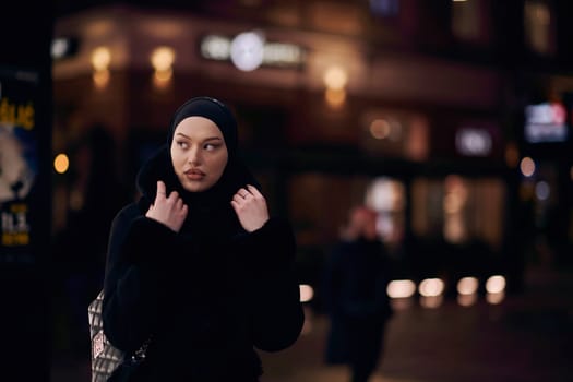 Young Muslim woman walking on urban city street on a cold winter night wearing hijab scarf veil a fashionable coat with bokeh city light in the background.