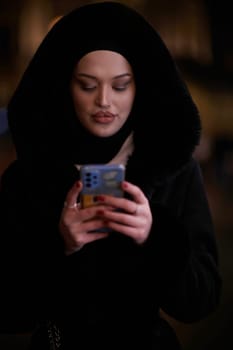 Young Muslim woman walking on urban city street on a cold winter night wearing hijab scarf veil a fashionable coat with bokeh city light in the background.