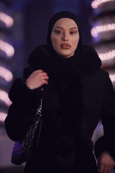 Young Muslim woman walking on urban city street on a cold winter night wearing hijab scarf veil a fashionable coat with bokeh city light in the background.