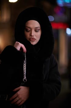 Young Muslim woman walking on urban city street on a cold winter night wearing hijab scarf veil a fashionable coat with bokeh city light in the background.