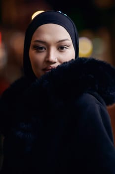 Young Muslim woman walking on urban city street on a cold winter night wearing hijab scarf veil a fashionable coat with bokeh city light in the background.