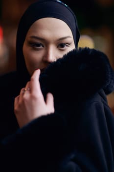 Young Muslim woman walking on urban city street on a cold winter night wearing hijab scarf veil a fashionable coat with bokeh city light in the background.