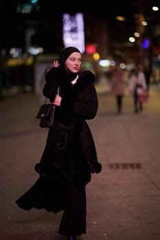 Young Muslim woman walking on urban city street on a cold winter night wearing hijab scarf veil a fashionable coat with bokeh city light in the background.