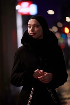 Young Muslim woman walking on urban city street on a cold winter night wearing hijab scarf veil a fashionable coat with bokeh city light in the background.