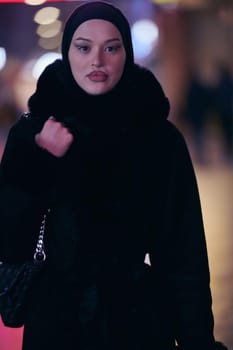 Young Muslim woman walking on urban city street on a cold winter night wearing hijab scarf veil a fashionable coat with bokeh city light in the background.