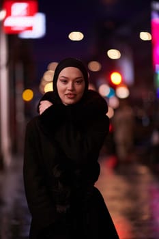 Young Muslim woman walking on urban city street on a cold winter night wearing hijab scarf veil a fashionable coat with bokeh city light in the background.