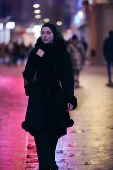 Young Muslim woman walking on urban city street on a cold winter night wearing hijab scarf veil a fashionable coat with bokeh city light in the background.