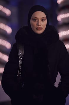 Young Muslim woman walking on urban city street on a cold winter night wearing hijab scarf veil a fashionable coat with bokeh city light in the background.