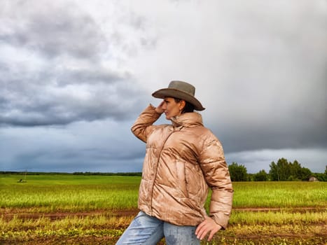 An adult girl looking like a cowboy in a hat in a field and with a stormy sky with clouds takes pictures of rainbow and takes selfie in the rain. Woman having fun outdoors on rural and rustic nature