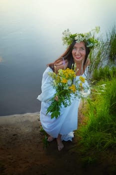 Adult mature brunette woman in a white dress, sundress and a wreath of flowers in summer by the water of river or lake in evening at sunset. Celebration of the Slavic pagan holiday of Ivan Kupala