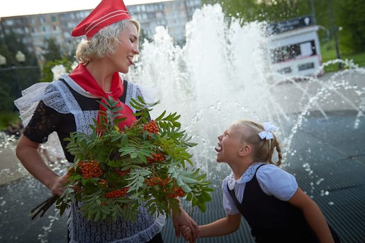 Young and adult schoolgirl on September with flowers having fun near water of fontain. Generations of schoolchildren, pioneer of USSR and October girl in modern uniform of Russia. Mom and daughter