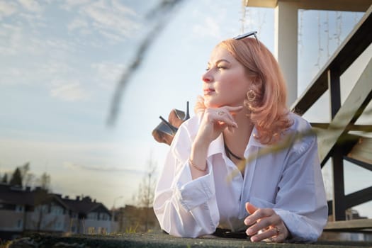 Portrait of Beautiful girl with red hair in white shirt in open wooden pavillion in village or small town. Young slender woman and sky with clouds on background on autumn, spring or summer evening