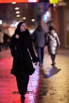 Young Muslim woman walking on urban city street on a cold winter night wearing hijab scarf veil a fashionable coat with bokeh city light in the background.