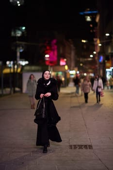 Young Muslim woman walking on urban city street on a cold winter night wearing hijab scarf veil a fashionable coat with bokeh city light in the background.