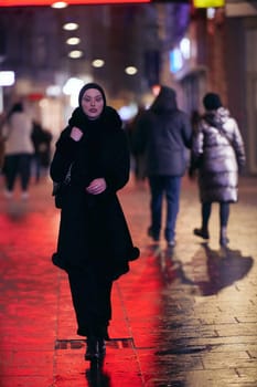 Young Muslim woman walking on urban city street on a cold winter night wearing hijab scarf veil a fashionable coat with bokeh city light in the background.