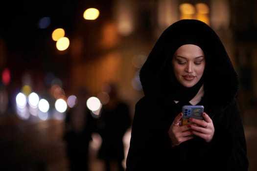 Young Muslim woman walking on urban city street on a cold winter night wearing hijab scarf veil a fashionable coat with bokeh city light in the background.