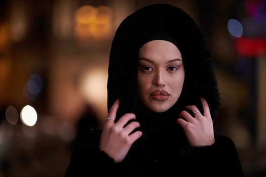 Young Muslim woman walking on urban city street on a cold winter night wearing hijab scarf veil a fashionable coat with bokeh city light in the background.