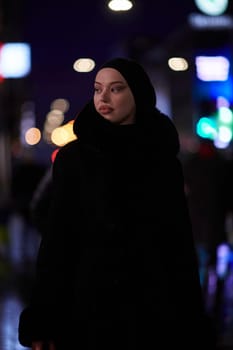 Young Muslim woman walking on urban city street on a cold winter night wearing hijab scarf veil a fashionable coat with bokeh city light in the background.