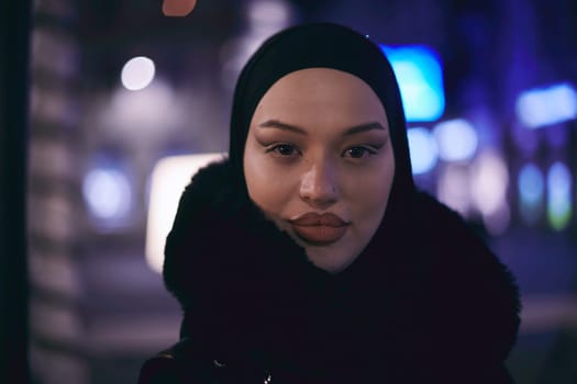 Young Muslim woman walking on urban city street on a cold winter night wearing hijab scarf veil a fashionable coat with bokeh city light in the background.