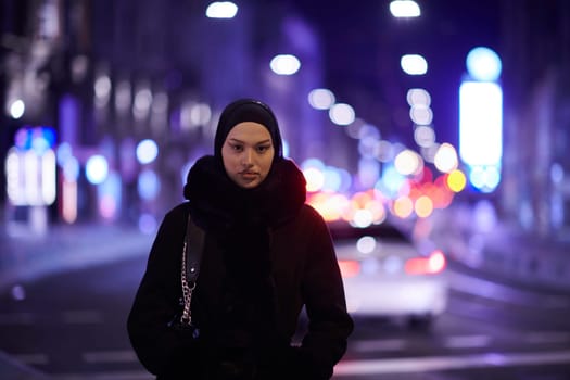 Young Muslim woman walking on urban city street on a cold winter night wearing hijab scarf veil a fashionable coat with bokeh city light in the background.