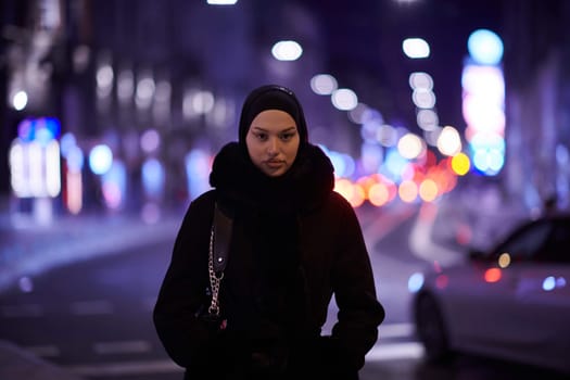 Young Muslim woman walking on urban city street on a cold winter night wearing hijab scarf veil a fashionable coat with bokeh city light in the background.