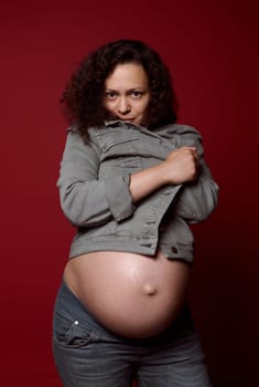 Attractive African American young pregnant woman with naked belly, wearing stylish casual denim blazer and jeans, isolated on red studio background. Pregnancy 36 week. Maternity lifestyle. Studio shot