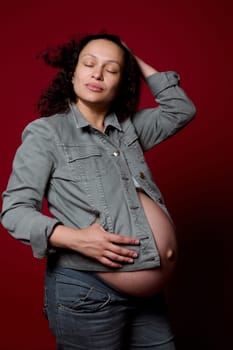 Beautiful multi-ethnic young pregnant woman, expectant mother with naked belly, wearing stylish casual denim blazer and jeans, posing over isolated red color studio background