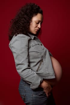 Side portrait of a pregnant woman, expectant mother in casual denim clothes, posing with her eyes closed over red isolated studio background. Pregnancy 36 week. Childbearing. Maternity lifestyle