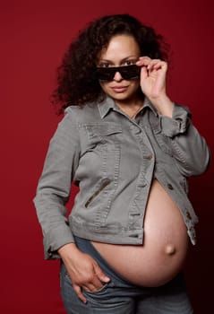 Pregnant woman dressed in casual jeans clothes, trendy sunglasses, posing with naked belly, isolated over red studio background. Childbearing. Pregnancy., Maternity fashion and lifestyle