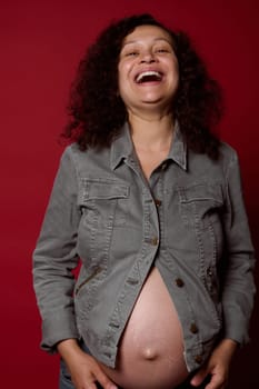 Happy curly haired multi-ethnic pregnant woman with naked belly, wearing stylish casual denim blazer and jeans, laughing looking at camera