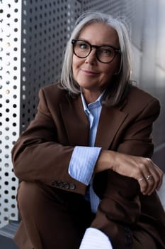 60 years old successful slender gray-haired business woman dressed in a stylish brown jacket is resting standing against the backdrop of a building wall.