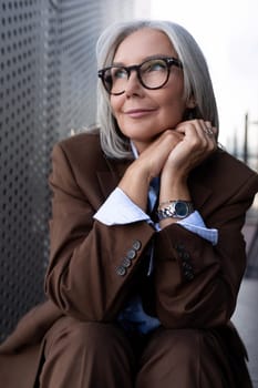 a beautiful gray-haired successful mature business woman in glasses dressed in a respectable way spends a coffee break on the street near the business center.