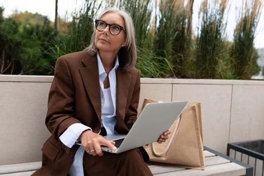 well-groomed charming entrepreneur woman of mature years works remotely sitting on a bench with a laptop.