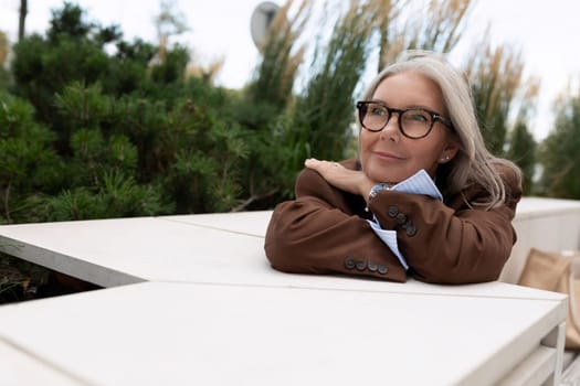 a mature woman with gray hair in a brown jacket is sitting at a table on the terrace of a cafe waiting for a partner.