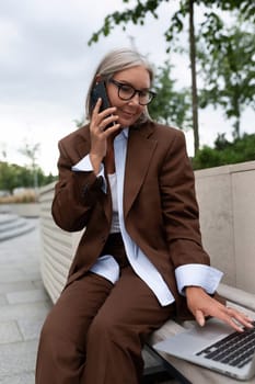 pretty gray-haired retired woman dressed in an elegant suit talking on the phone while walking down the street.