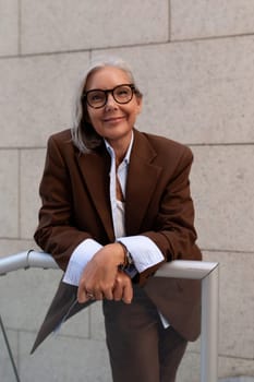 portrait of a model of a slender gray-haired woman of mature years dressed in an elegant brown suit against the background of a wall of a building.