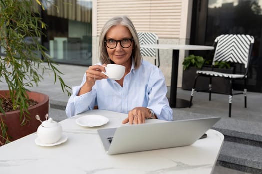 business portrait of a confident successful 60 year old gray-haired woman in glasses sitting during a business lunch on the terrace.
