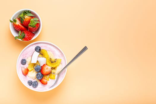 Healthy breakfast or dessert yogurt bowl with fresh banana, strawberry, blueberry, cocos, kiwi top view on minimal pastel paper background with spoon. Copy space